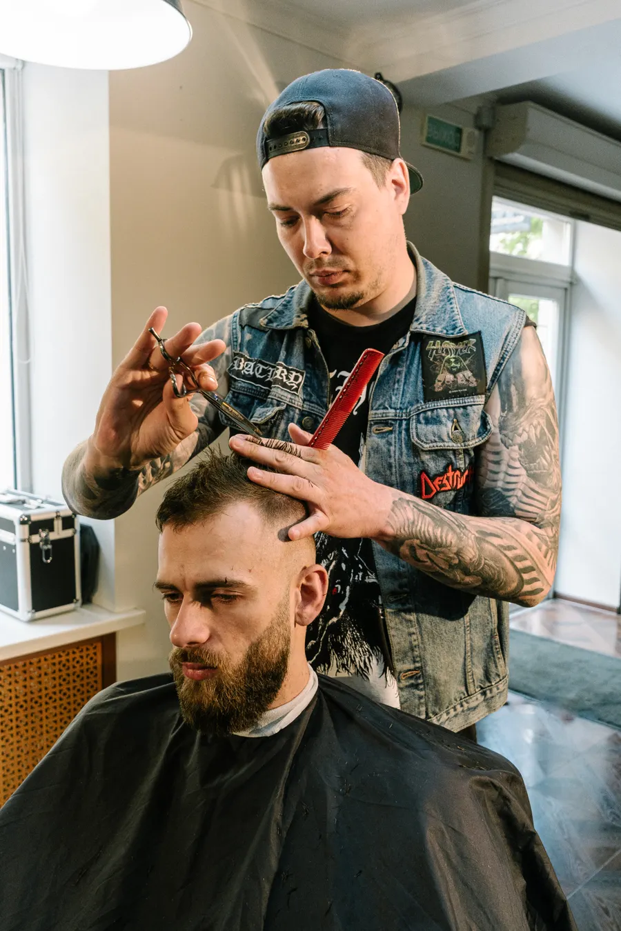 Jake Sullivan, tattooed barber in a denim vest, cutting hair with scissors and a comb
