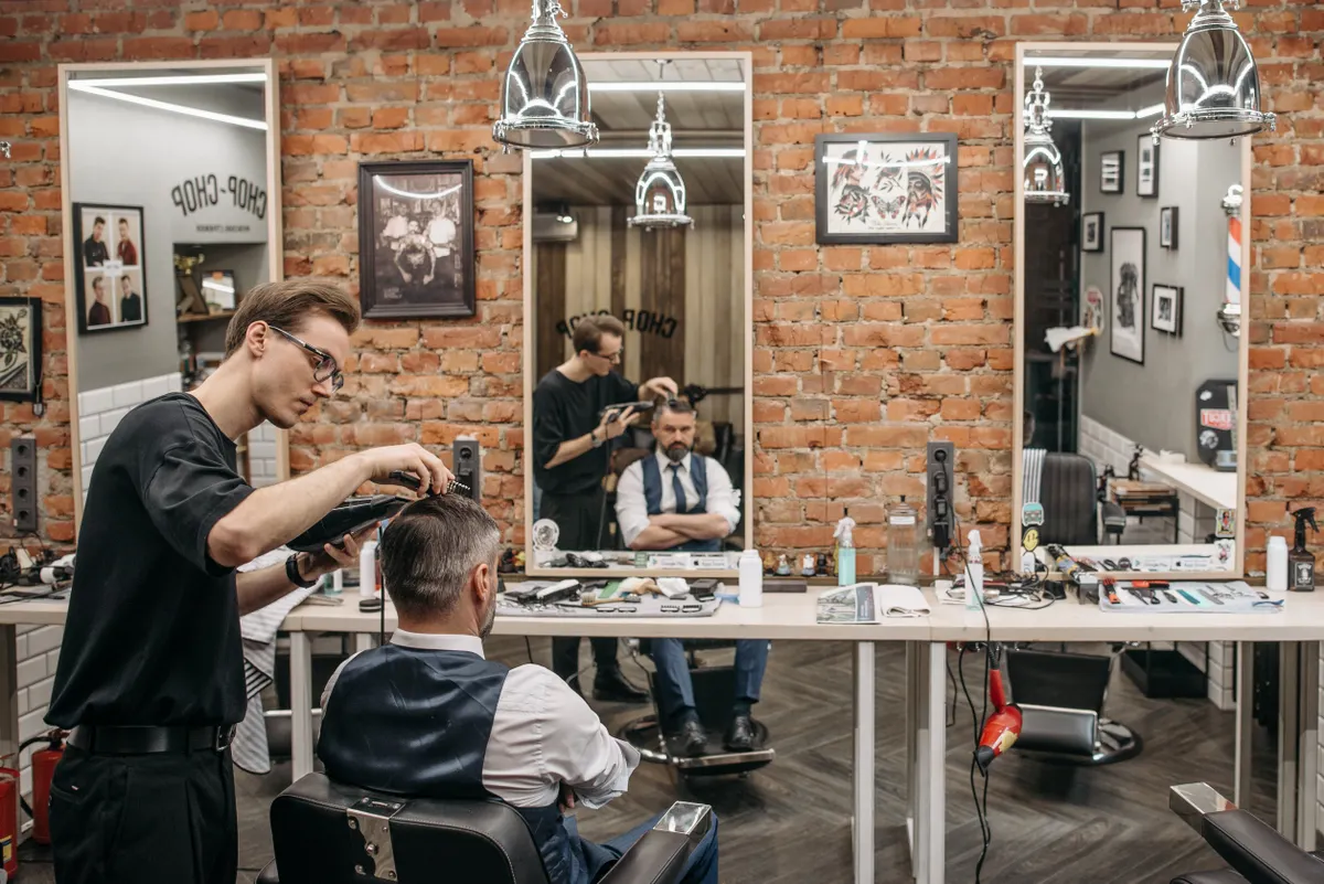 Owen Mitchell in glasses cutting a client’s hair in a brick-walled barbershop